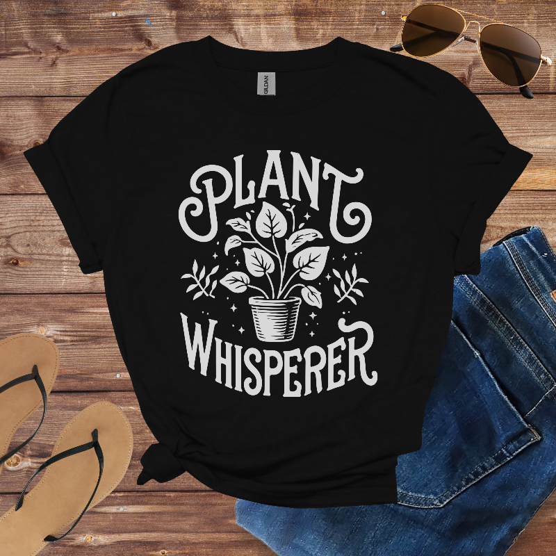 Black t-shirt with 'Plant Whisperer' design on a wooden surface with sunglasses, sandals, and jeans.