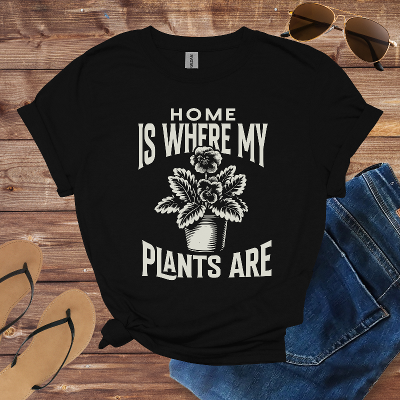 Black t-shirt with 'Home is where my plants are' design on a wooden surface with sunglasses, flip-flops, and jeans.