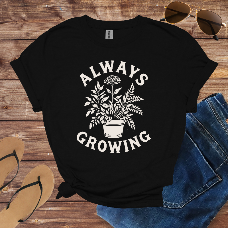 Black t-shirt with 'Always Growing' graphic on a wooden surface with sunglasses and jeans.