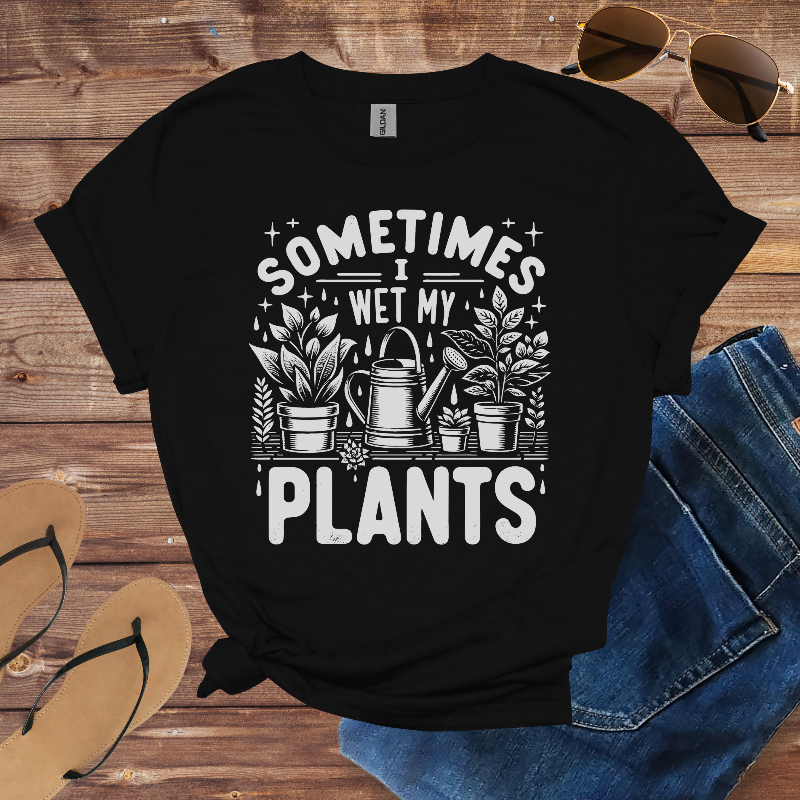 Black t-shirt with 'Sometimes I Wet My Plants' design on a wooden surface with sunglasses, flip-flops, and jeans.