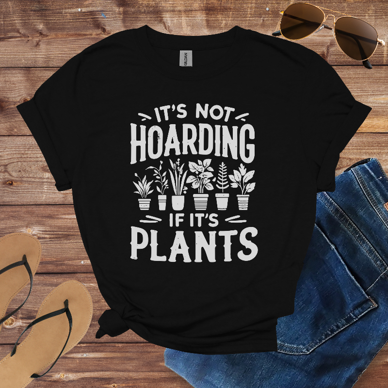 Black t-shirt with 'It's not hoarding if it's plants' text and plant illustrations on a wooden surface with sunglasses and jeans.