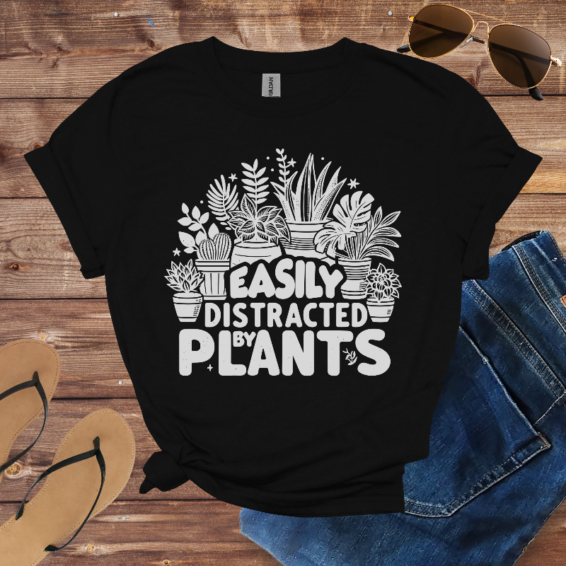 Black t-shirt with 'Easily Distracted by Plants' design on a wooden surface with sunglasses, jeans, and sandals.