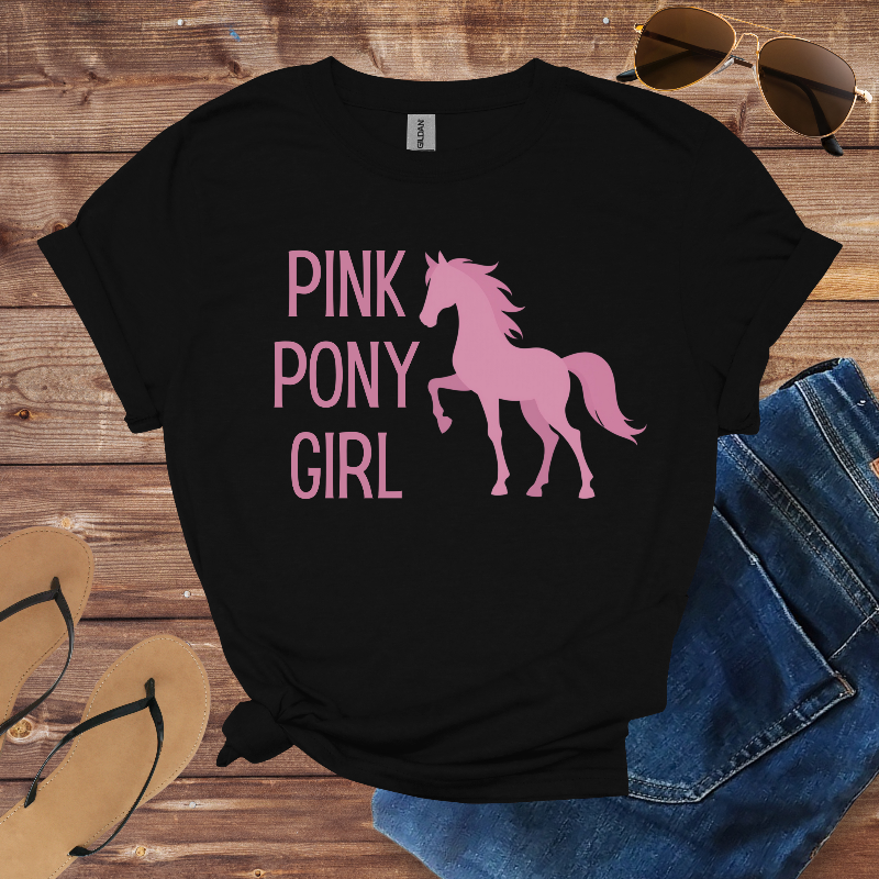 Black t-shirt with pink pony graphic and text on a wooden surface with sunglasses, jeans, and flip-flops.