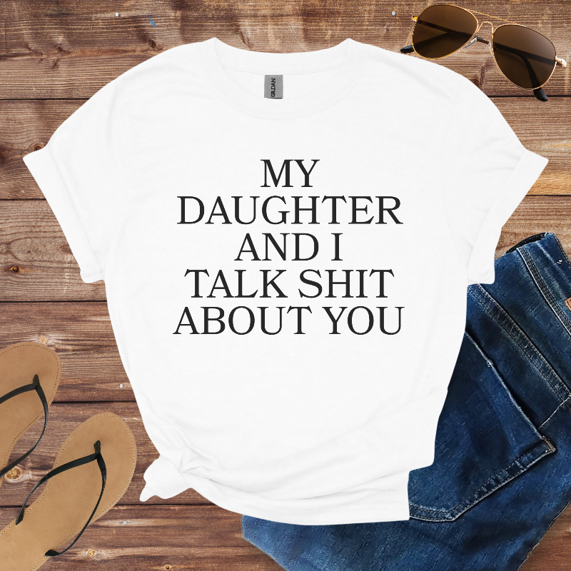 White t-shirt with text 'My daughter and I talk shit about you' on a wooden surface with sunglasses, jeans, and flip-flops.