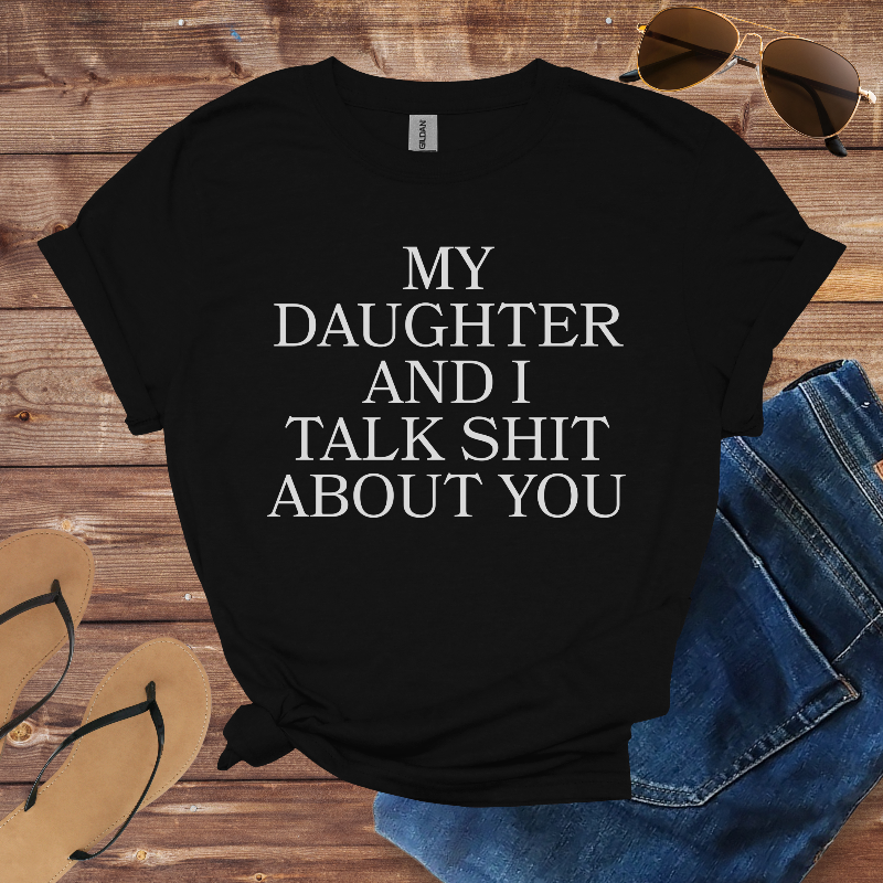 Black t-shirt with text 'My daughter and I talk shit about you' on a wooden surface with sunglasses, jeans, and flip-flops.