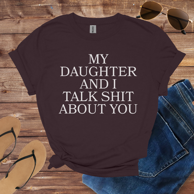 Brown t-shirt with text 'My daughter and I talk shit about you' on a wooden surface with sunglasses, jeans, and sandals.