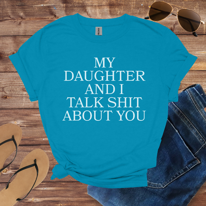 Blue t-shirt with text 'My daughter and I talk shit about you' on a wooden surface with sunglasses, jeans, and sandals.