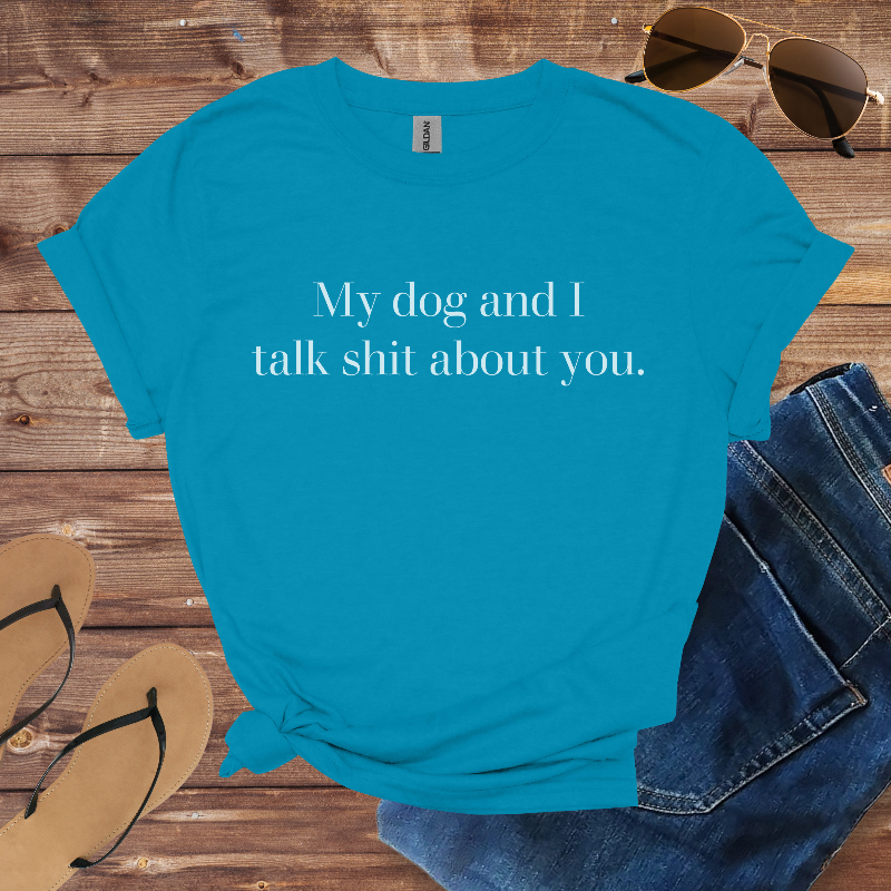 Blue t-shirt with text 'My dog and I talk shit about you' on a wooden surface with sunglasses, jeans, and flip-flops.