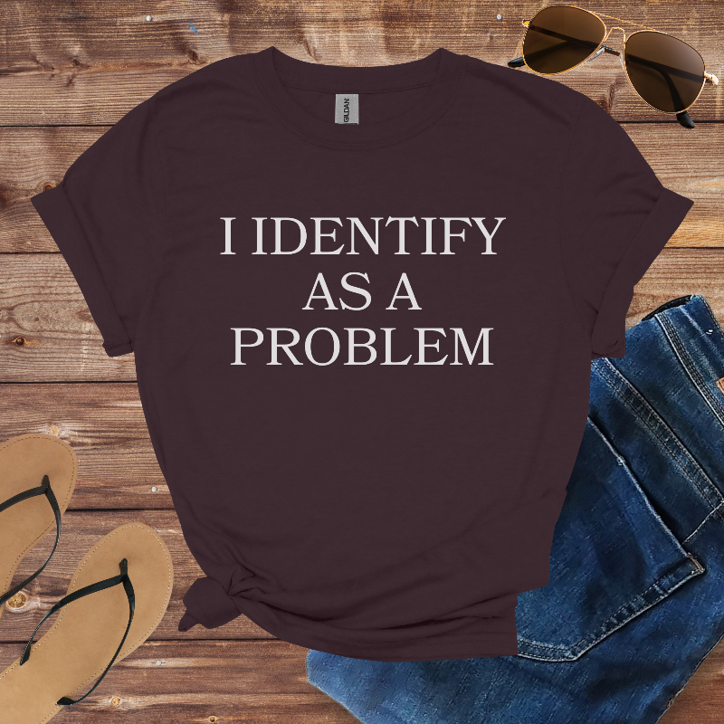 Maroon t-shirt with 'I IDENTIFY AS A PROBLEM' text on a wooden surface with sunglasses, jeans, and flip-flops.