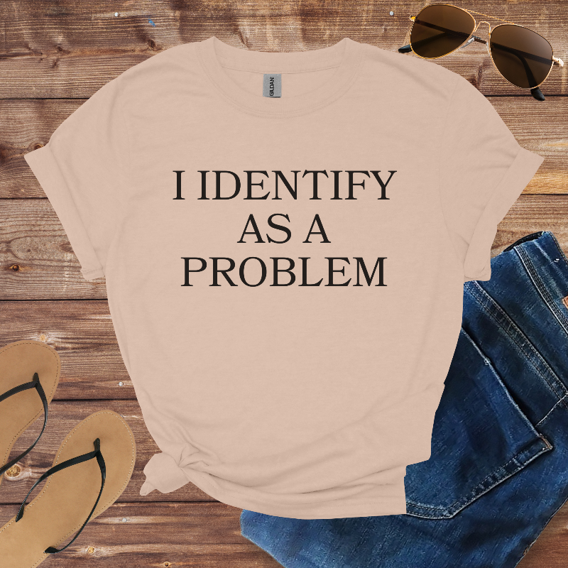 Beige t-shirt with 'I IDENTIFY AS A PROBLEM' text on a wooden surface with sunglasses, jeans, and sandals.