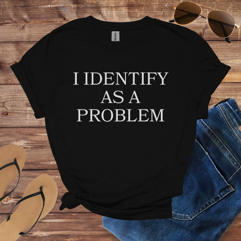 Black t-shirt with 'I IDENTIFY AS A PROBLEM' text on a wooden surface with sunglasses, jeans, and flip-flops.
