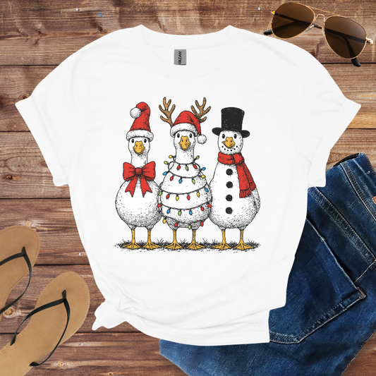 White t-shirt with Christmas-themed design of three ducks on a wooden surface with sunglasses and jeans.