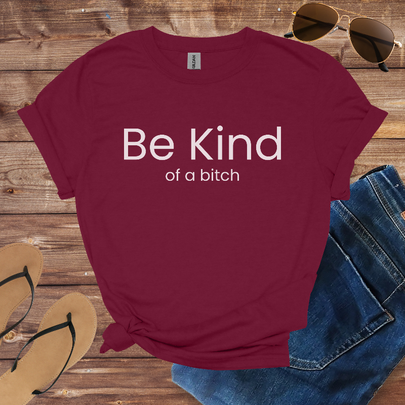 Maroon t-shirt with 'Be Kind of a bitch' text on a wooden surface with sunglasses, jeans, and sandals.