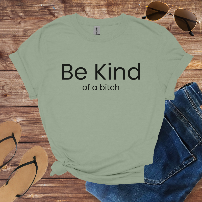 Green t-shirt with 'Be Kind of a bitch' text on a wooden surface with sunglasses, jeans, and sandals.