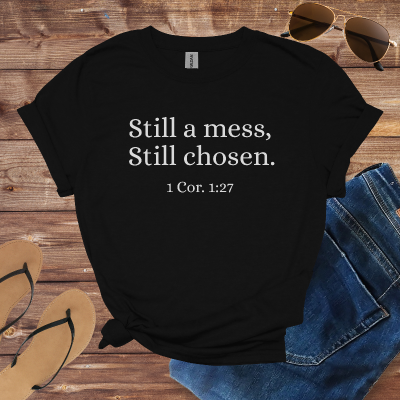 Black t-shirt with 'Still a mess, Still chosen. 1 Cor. 1:27' text on a wooden surface with sunglasses and jeans.