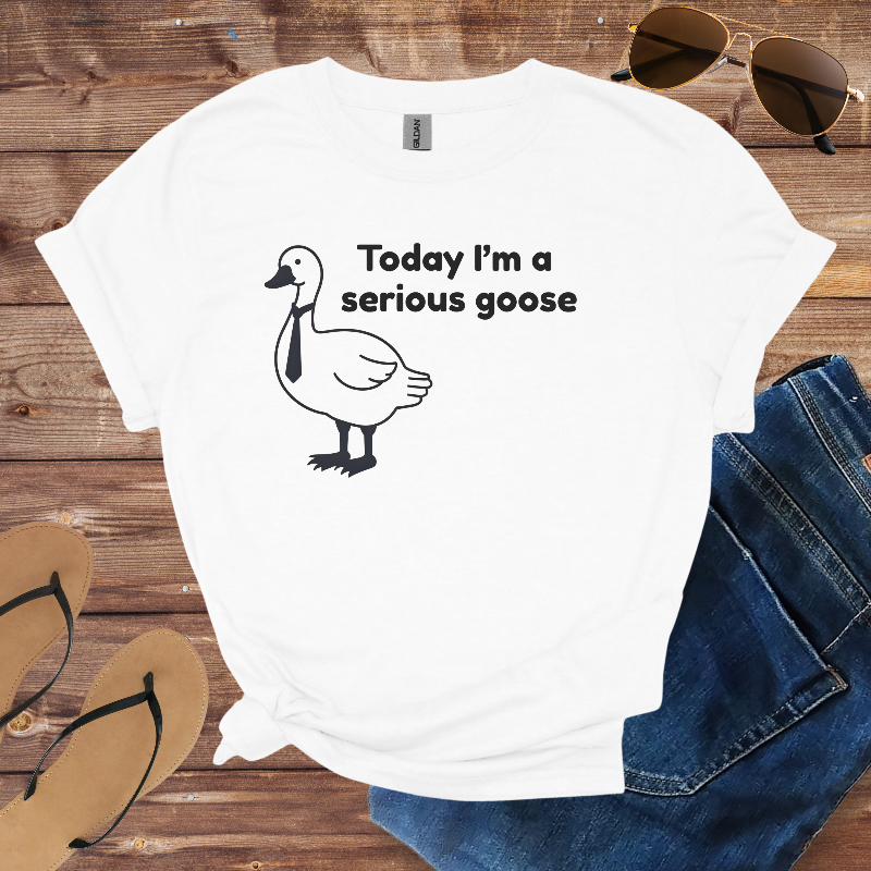 White t-shirt with a goose graphic and text on a wooden surface with sunglasses, jeans, and flip-flops.