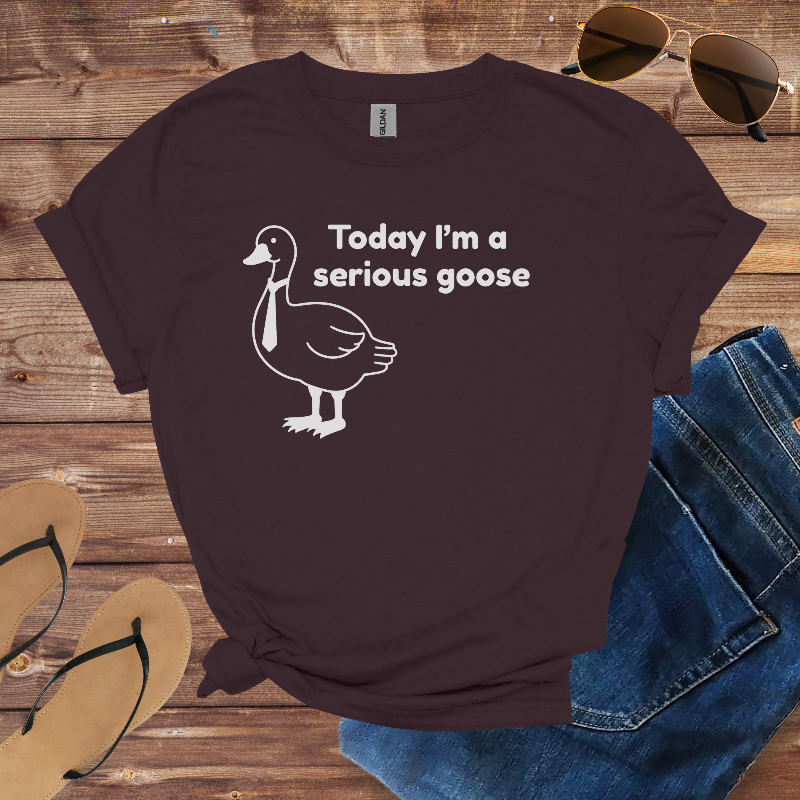 Brown t-shirt with a goose graphic and text on a wooden surface with sunglasses, flip-flops, and jeans.