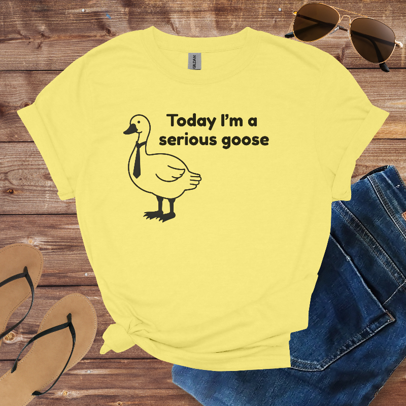 Yellow t-shirt with a goose graphic and text on a wooden surface with sunglasses, jeans, and sandals.