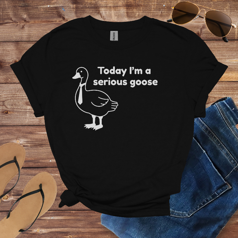Black t-shirt with a goose graphic and text on a wooden surface with sunglasses, jeans, and sandals.