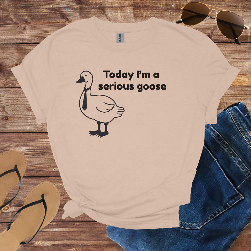 Beige t-shirt with a goose graphic and text on a wooden surface with sunglasses, jeans, and flip-flops.