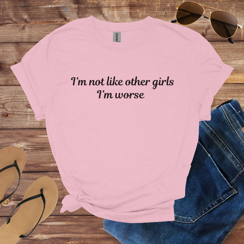 Pink t-shirt with text 'I'm not like other girls I'm worse' on a wooden surface with sunglasses, jeans, and flip-flops.