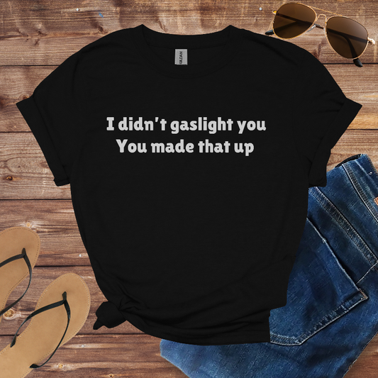 Black t-shirt with text 'I didn't gaslight you You made that up' on a wooden surface with sunglasses, jeans, and flip-flops.