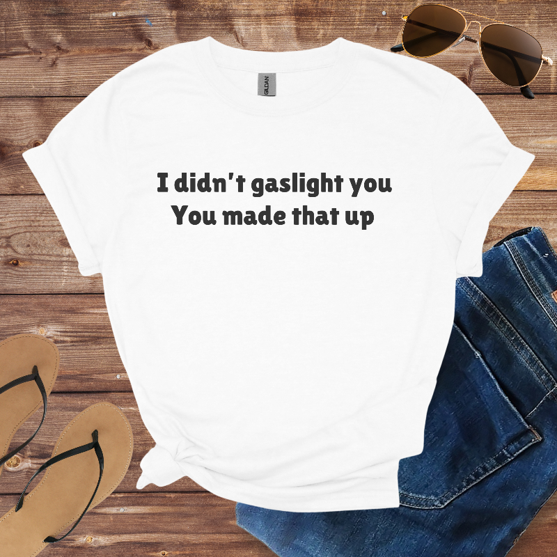 White t-shirt with text 'I didn't gaslight you You made that up' on a wooden surface with sunglasses, jeans, and flip-flops.