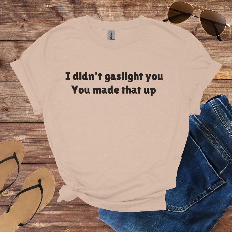 Beige t-shirt with text 'I didn't gaslight you You made that up' on a wooden surface with sunglasses, jeans, and flip-flops.
