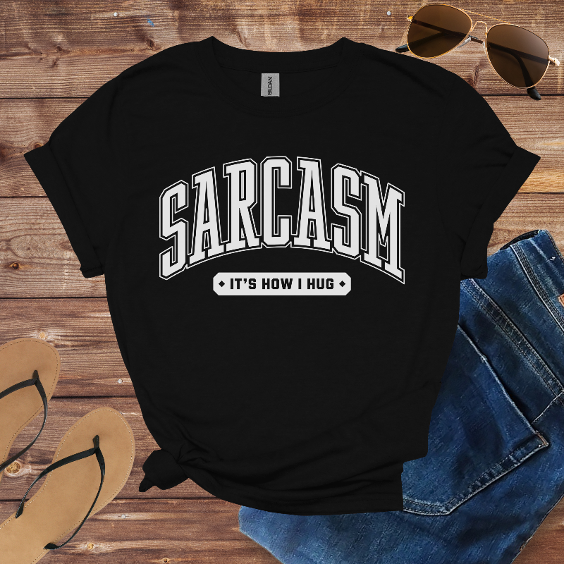 Black t-shirt with 'SARCASM IT'S HOW I HUG' text on a wooden surface with sunglasses and jeans.