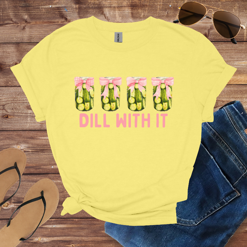Yellow t-shirt with 'Dill With It' text and pickle graphic on a wooden surface with sunglasses, jeans, and sandals.