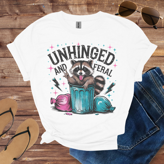 White t-shirt with raccoon graphic and text 'Unhinged and Feral' on a wooden surface with sunglasses, jeans, and sandals.