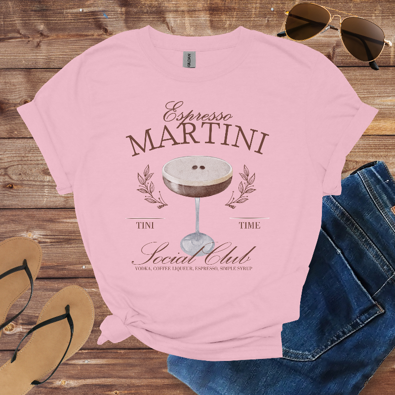 Pink t-shirt with 'Espresso Martini' design on a wooden surface with sunglasses, jeans, and flip-flops.