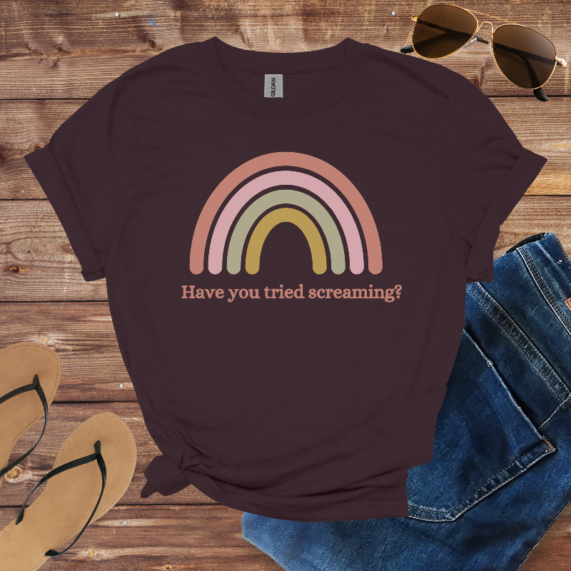 Brown t-shirt with rainbow design and text on a wooden surface with sunglasses, jeans, and sandals.