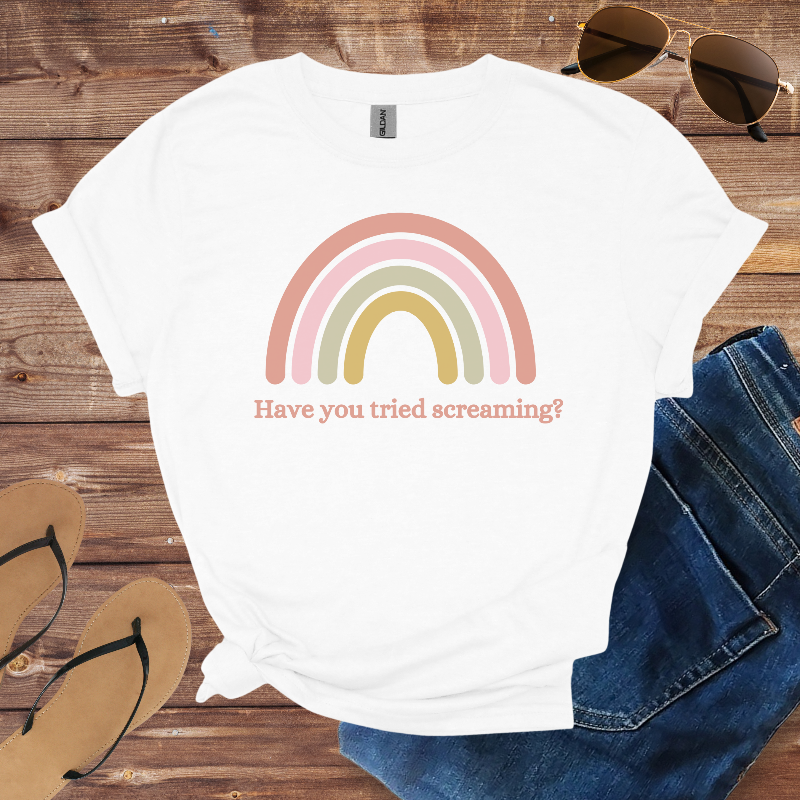 White t-shirt with rainbow design and text on a wooden surface with sunglasses, jeans, and sandals.