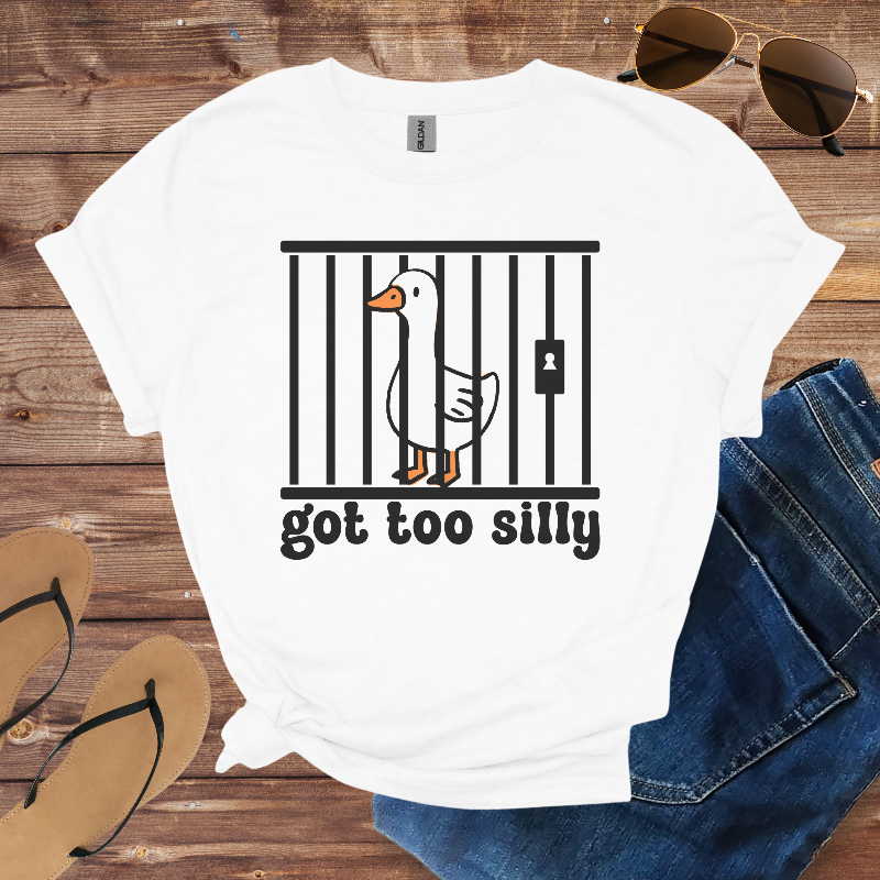 White t-shirt with a graphic of a duck in jail and 'got too silly' text, placed on a wooden surface with sunglasses, jeans, and flip-flops.