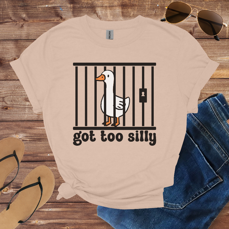 Beige t-shirt with a goose graphic and text 'got too silly' on a wooden surface with sunglasses, jeans, and sandals.