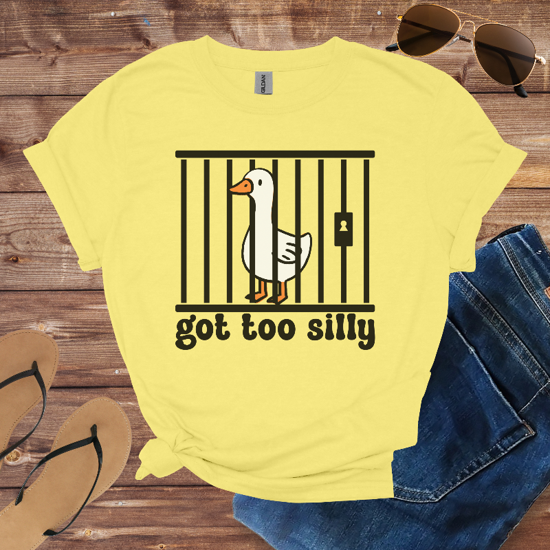 Yellow t-shirt with a goose graphic and text 'got too silly' on a wooden surface with sunglasses, jeans, and sandals.