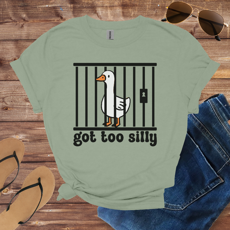 Green t-shirt with a goose graphic and text 'got too silly' on a wooden surface with sunglasses, jeans, and sandals.