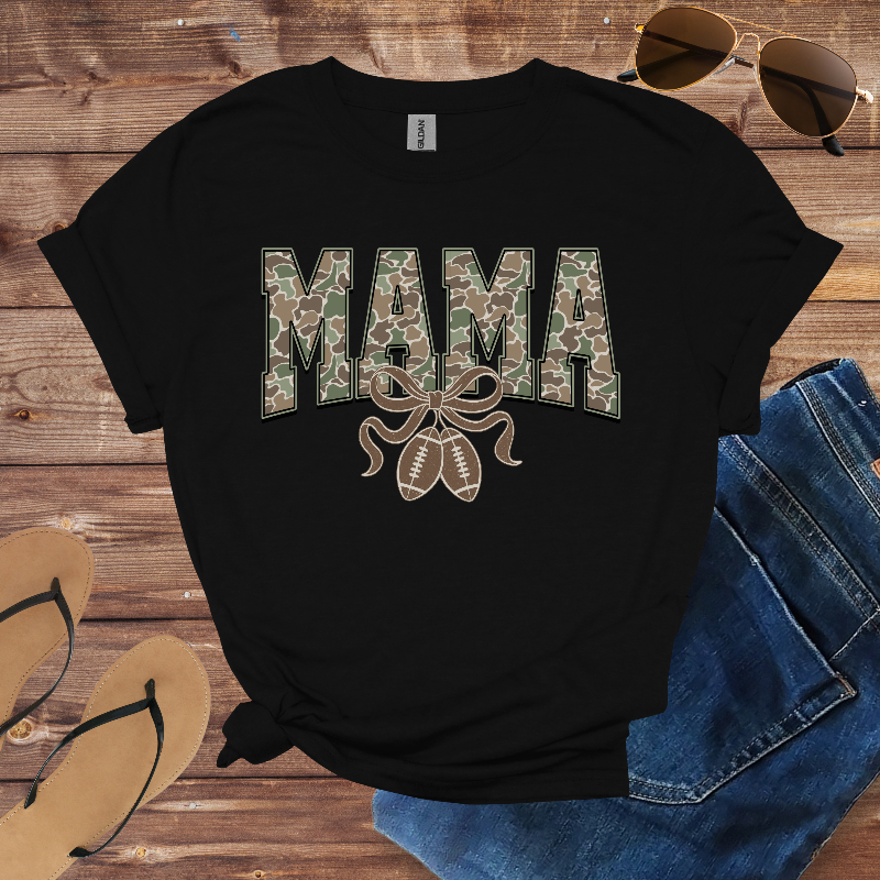 Black t-shirt with 'MAMA' in camouflage print on a wooden surface with sunglasses and jeans.