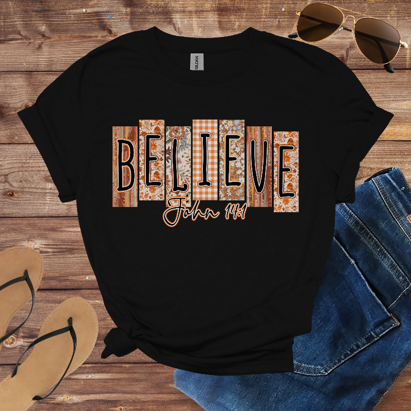 Black t-shirt with 'BELIEVE John 3:16' text on a wooden surface with sunglasses and jeans.