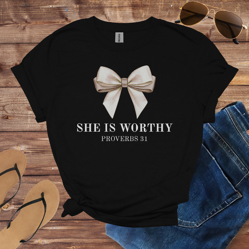 Black t-shirt with a bow design and 'SHE IS WORTHY Proverbs 31' text on a wooden surface with sunglasses and jeans.