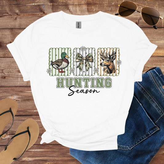 White t-shirt with hunting-themed design on a wooden surface with sunglasses, jeans, and flip-flops.
