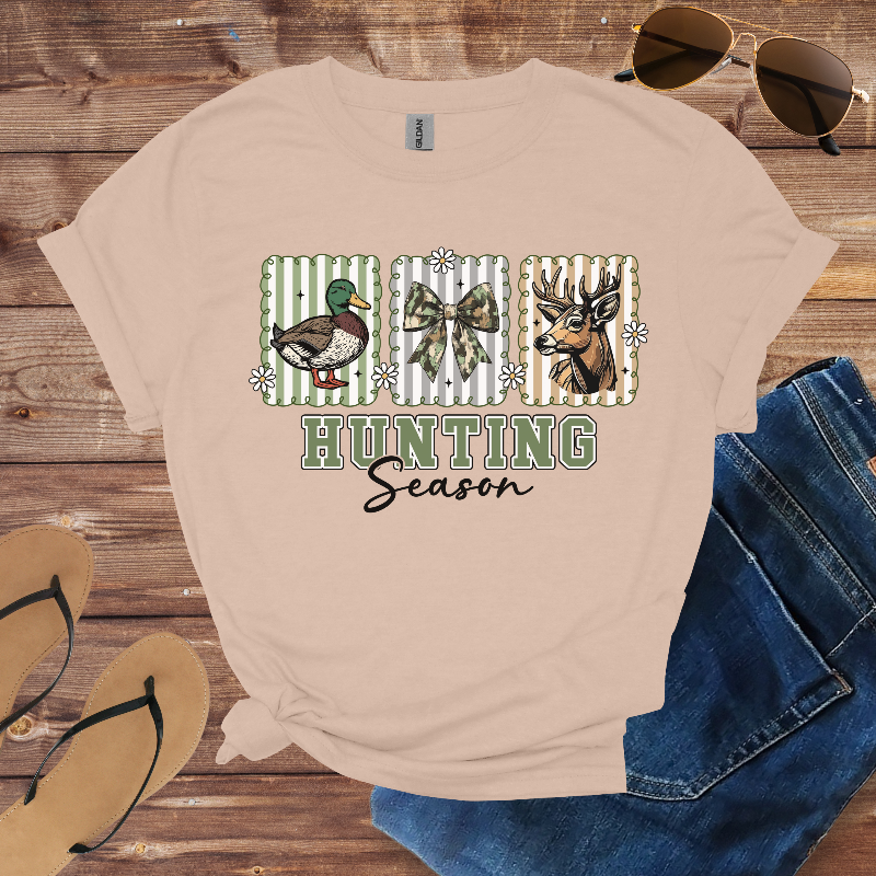 Beige t-shirt with hunting-themed design on a wooden surface with sunglasses and jeans.