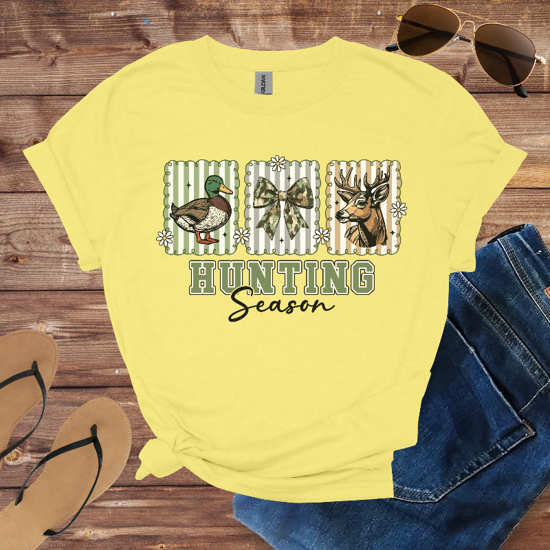 Yellow t-shirt with hunting-themed design on a wooden surface with sunglasses, jeans, and flip-flops.