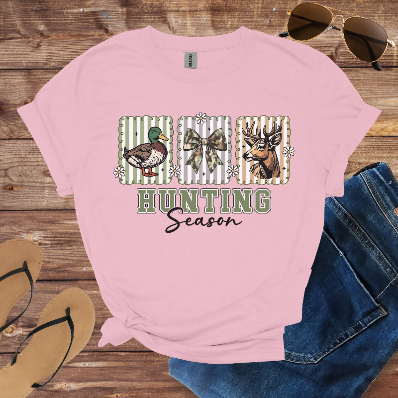Pink t-shirt with hunting-themed design on a wooden surface with sunglasses, jeans, and flip-flops.