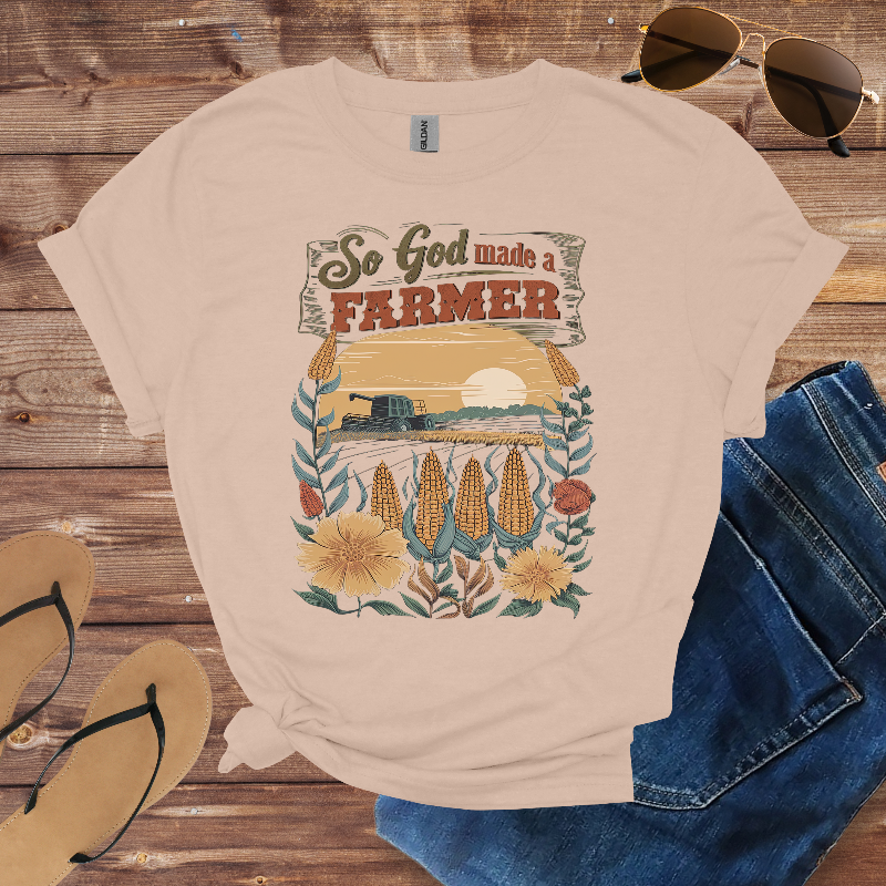 Beige t-shirt with 'So God made a farmer' graphic design on a wooden surface with sunglasses, jeans, and sandals.