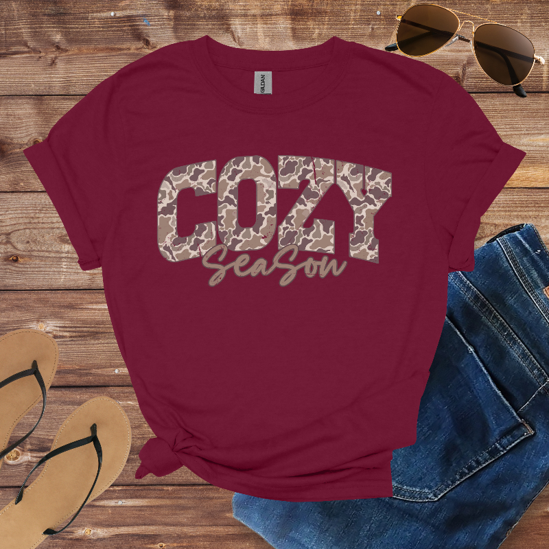 Maroon t-shirt with 'COZY Season' text on a wooden surface with sunglasses, jeans, and flip-flops.