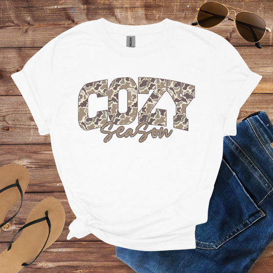 White t-shirt with 'Cozy Season' print on a wooden surface with sunglasses, jeans, and flip-flops.
