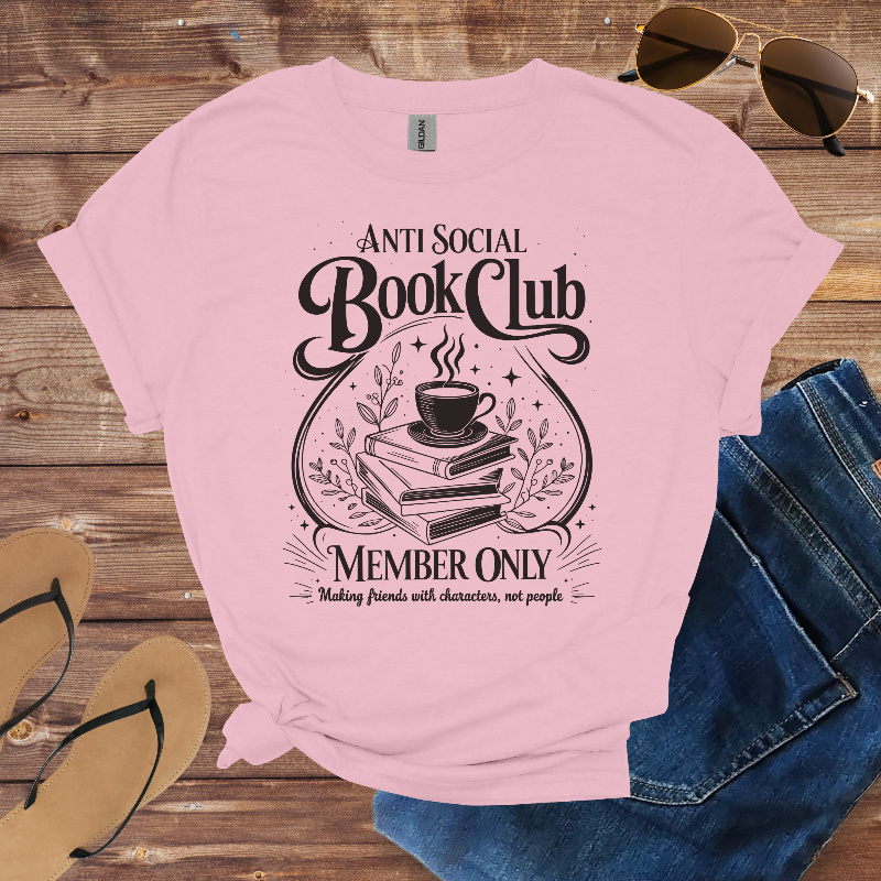 Pink t-shirt with 'Anti Social Book Club' design on a wooden surface with sunglasses, jeans, and sandals.