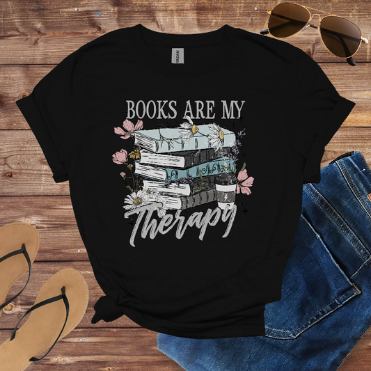 Black t-shirt with 'Books are my Therapy' design on a wooden surface with sunglasses, jeans, and sandals.